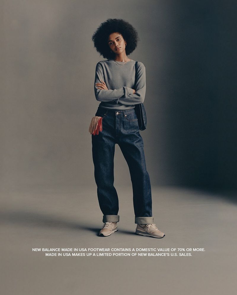 Fashion shot of woman with afro wearing grey sweater, jeans, and sneakers in a studio, with New Balance logo and branding.