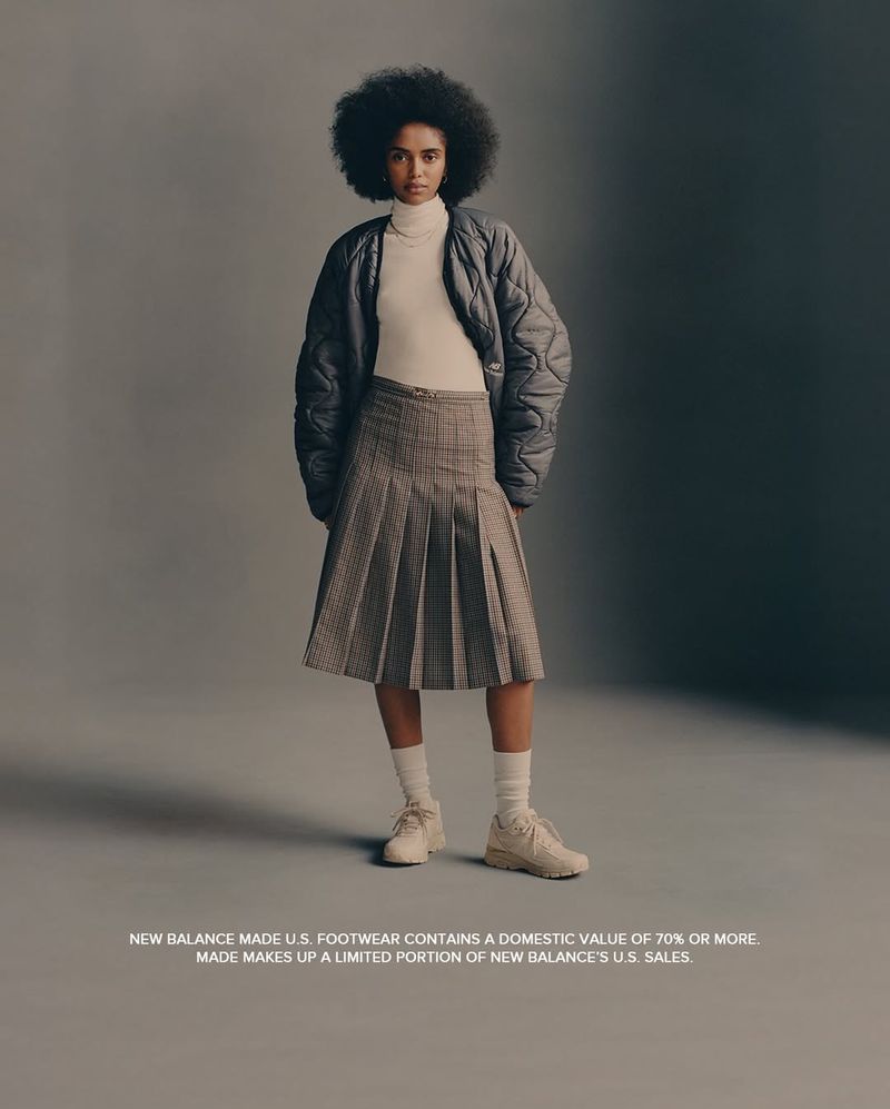 Full shot of a woman posing in a gray quilted jacket, beige turtleneck, pleated plaid skirt, white socks, and New Balance sneakers.