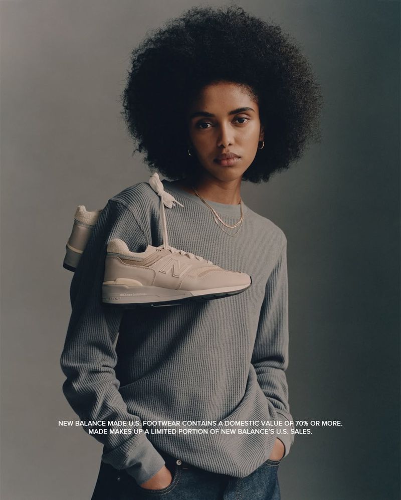 A young Black woman models New Balance sneakers. She is wearing a gray thermal and blue jeans in a studio setting, holding shoes.