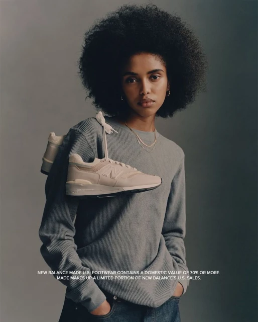 A young Black woman models New Balance sneakers. She is wearing a gray thermal and blue jeans in a studio setting, holding shoes.