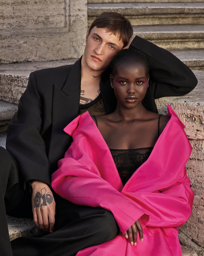 Fashion shot of an adult male with tattoos and an African woman in pink sitting on steps, wearing fashionable clothes in an outdoor setting.