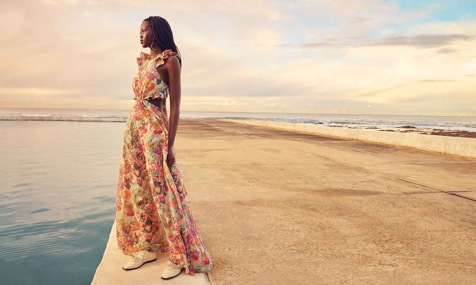 An adult female fashion model wears a floral-print maxi dress while posing on a concrete platform with an ocean and cloudy sky background.