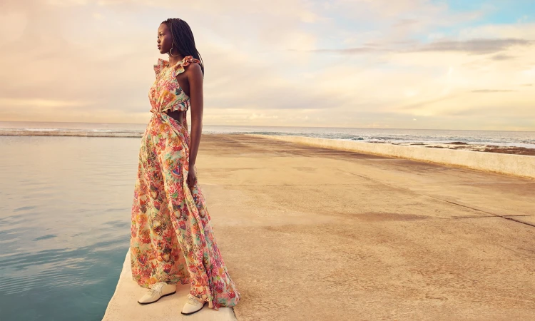 An adult female fashion model wears a floral-print maxi dress while posing on a concrete platform with an ocean and cloudy sky background.