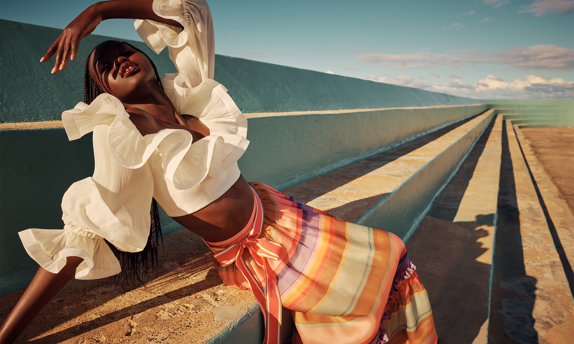 Adut Akech modeling Zimmermann Resort RTW collection, wearing ruffled white top and striped skirt on concrete bleachers.