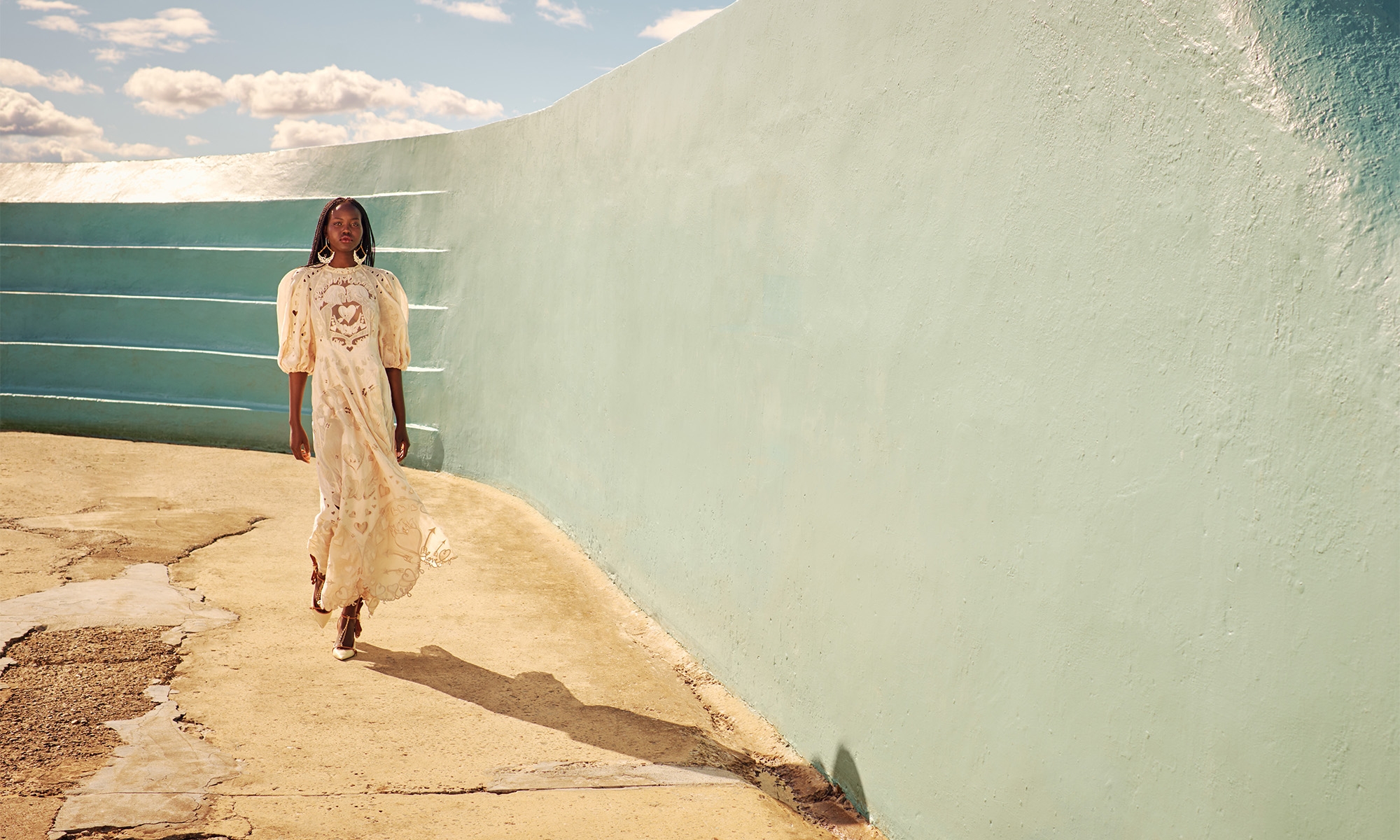 A striking fashion image features an African-American woman in a flowing dress, walking confidently in front of a light-blue wall and sky.