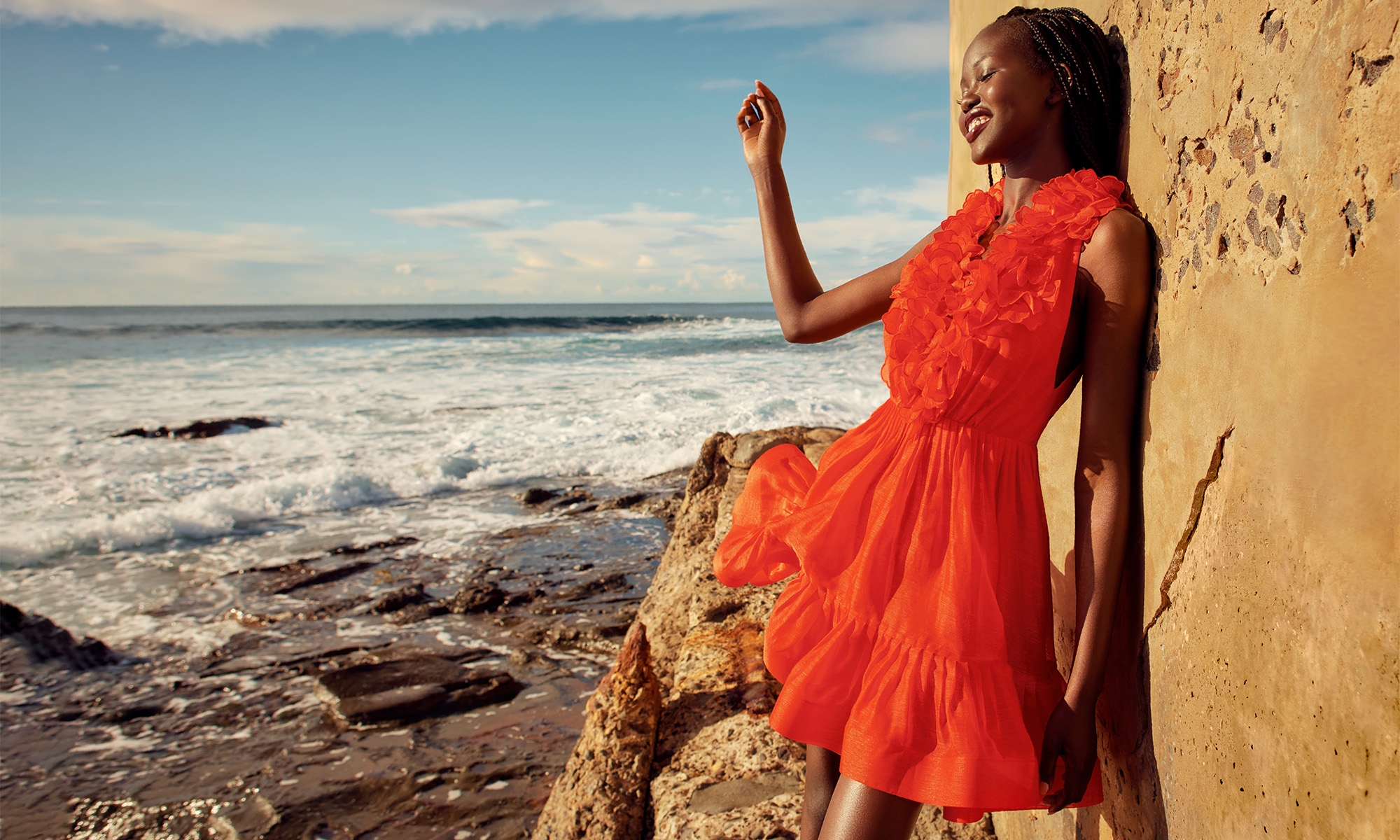 Adut Akech wearing a bright red ruffled dress leaning against a rocky stone wall by the ocean for Zimmermann Resort RTW.