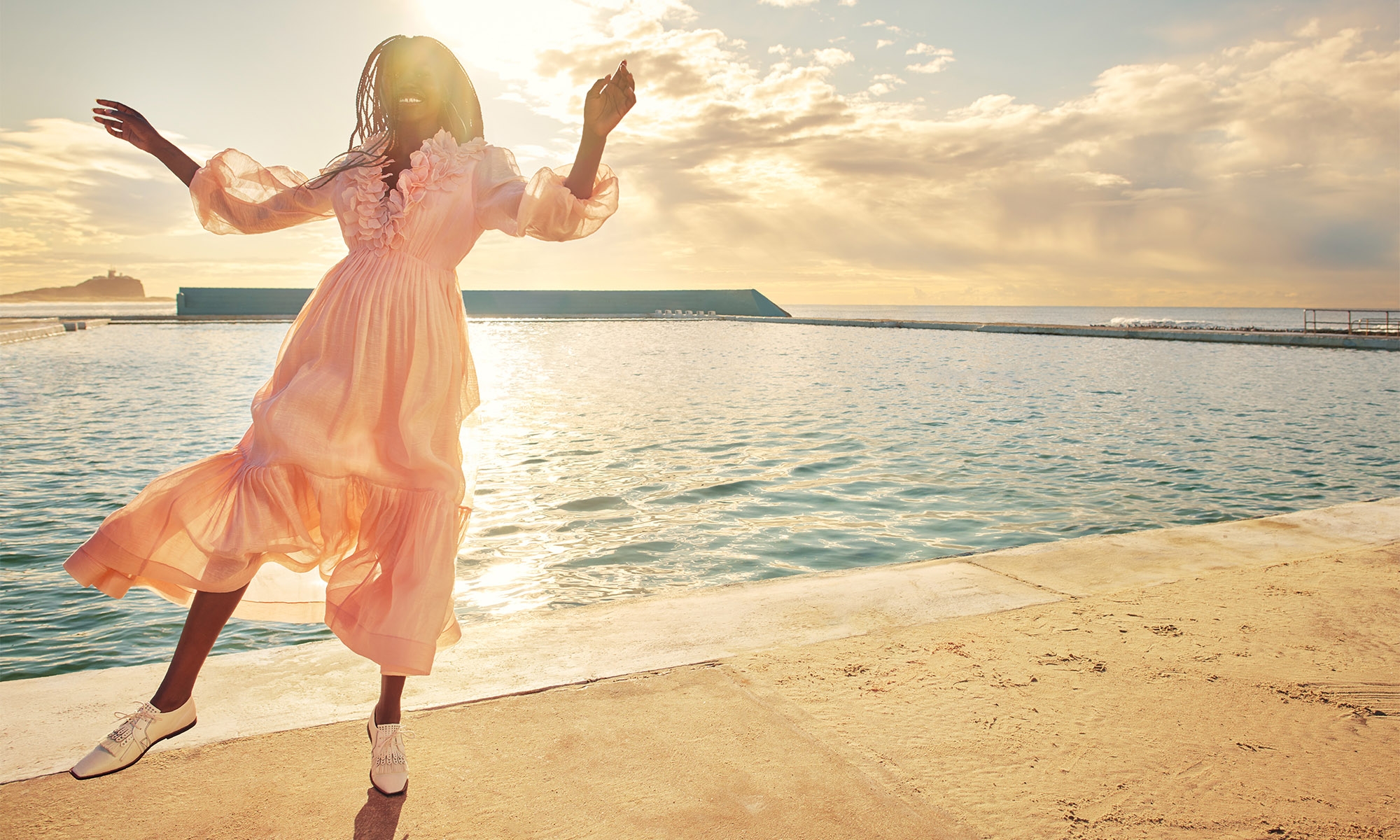 A woman in a pink dress stands near an ocean pool, with the sun casting a golden glow over the water and a distant rock formation.