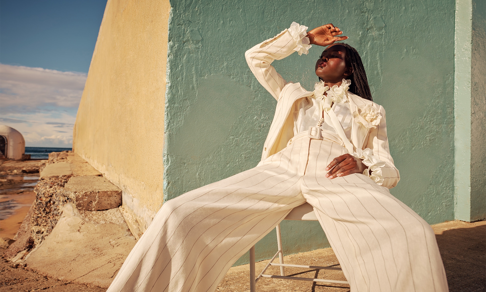 Fashion shot of an African American adult female in a pinstripe suit, posed on a chair against a geometric wall and ocean backdrop.