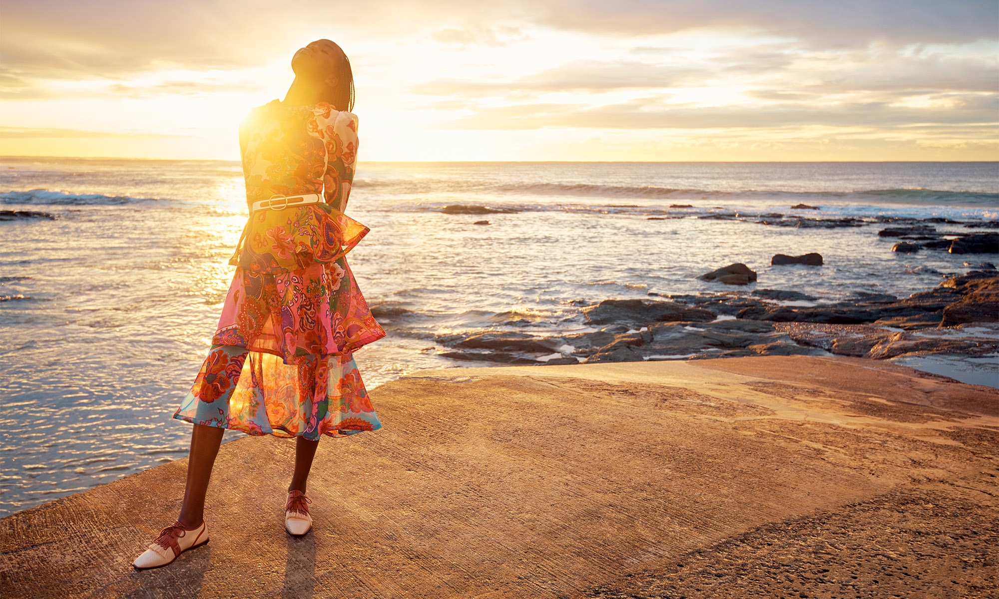 Woman in vibrant floral dress posing on a concrete pier at sunset over the ocean; golden light reflecting off the water.