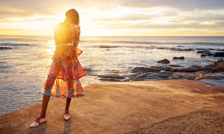 Woman in vibrant floral dress posing on a concrete pier at sunset over the ocean; golden light reflecting off the water.