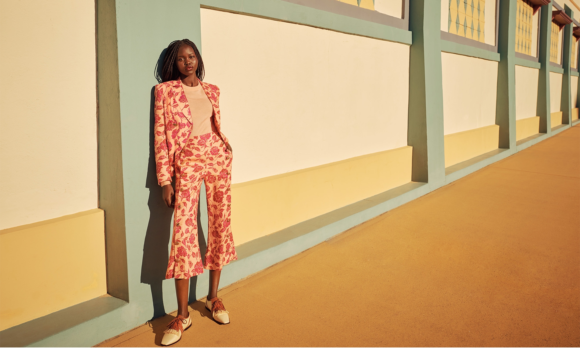 A young adult Black woman in a salmon-colored floral pantsuit stands against a segmented wall bathed in sunlight, casting shadows on the setting.