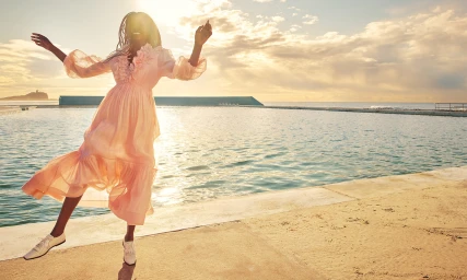 Model Adut Akech in a sheer pink dress dancing by an outdoor pool at sunset for Zimmermann Resort RTW.