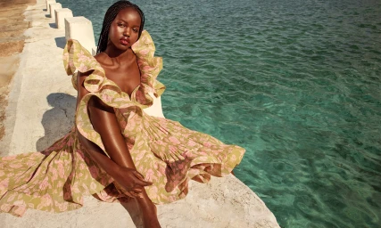 Model Adut Akech wearing a floral ruffled dress seated on a concrete seawall beside turquoise ocean water