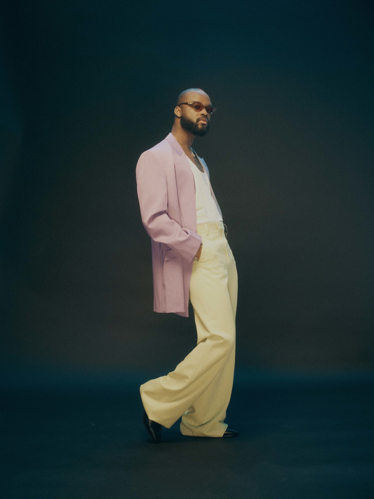 Stylish man in light pink suit, cream pants, and sunglasses poses in a studio with a confident expression, showcasing modern fashion.