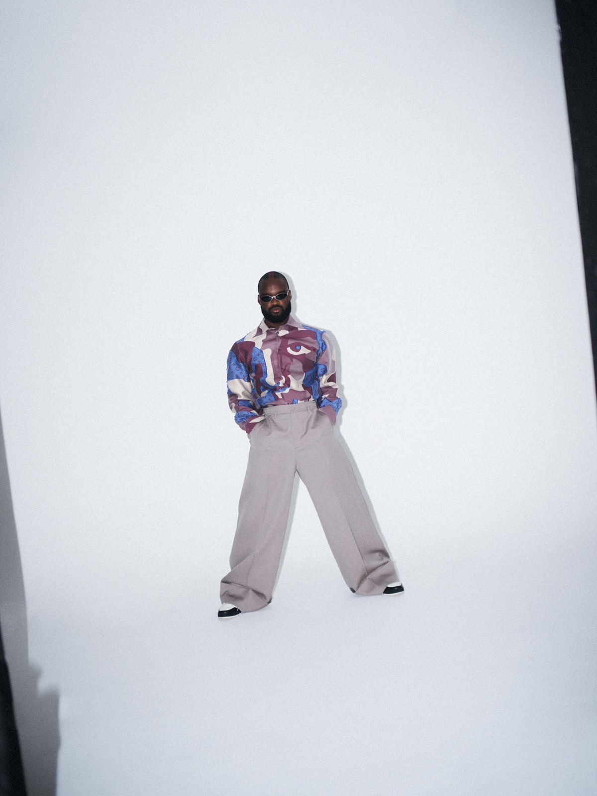 Man wearing a patterned shirt and wide-cut grey trousers posing against a plain white studio background.