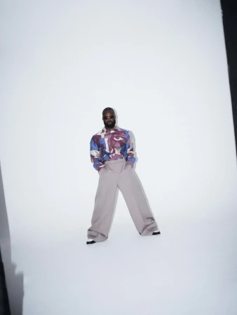 Man wearing a patterned shirt and wide-cut grey trousers posing against a plain white studio background.