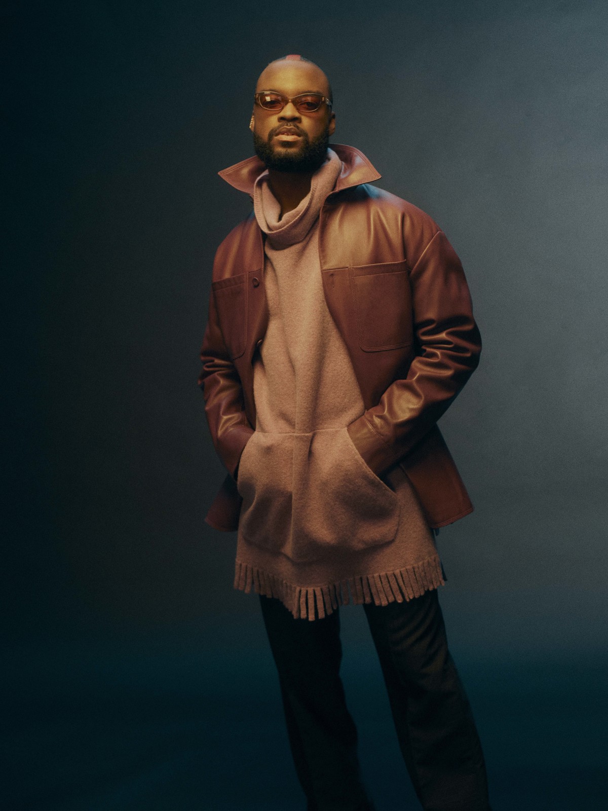 Portrait of an adult male wearing a brown jacket, beige sweater, and sunglasses posing in a studio setting with hands in pockets.