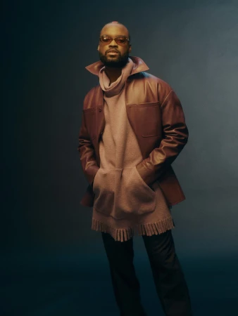 Portrait of an adult male wearing a brown jacket, beige sweater, and sunglasses posing in a studio setting with hands in pockets.