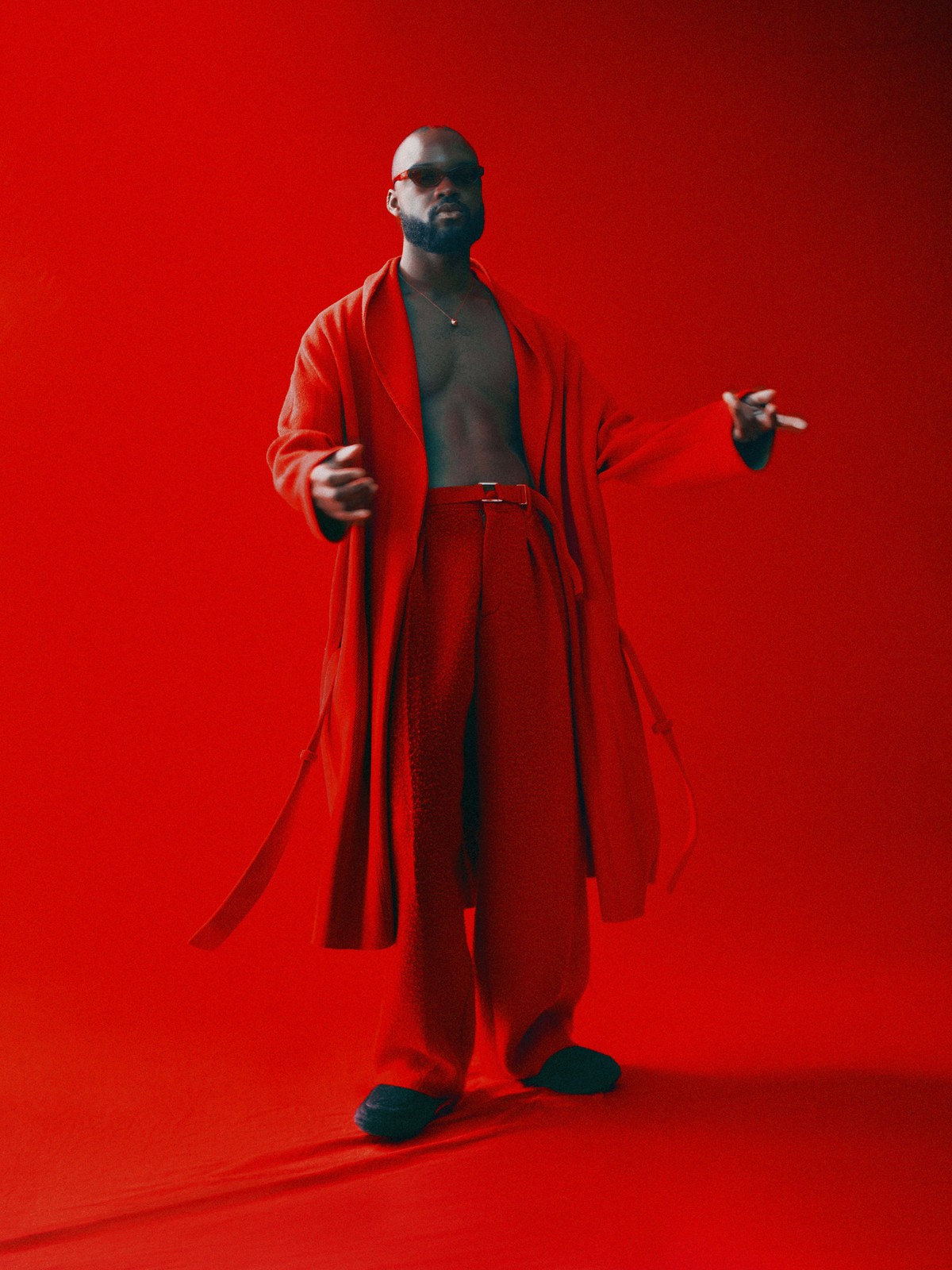 Full-length portrait of a bald Black man in a red robe and trousers against a solid red background in a studio setting.