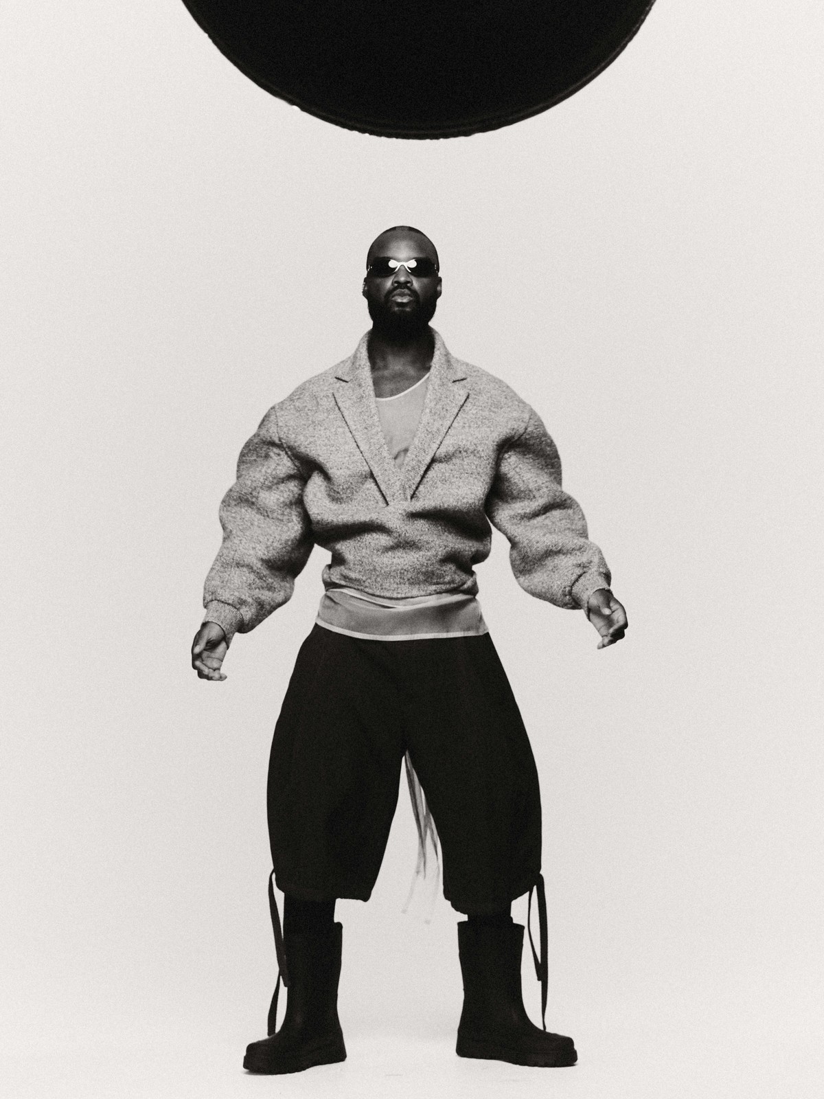 Black man wearing oversized grey blazer, dark shorts and black boots against a white studio background with a black disc overhead.