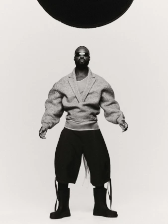 Black man wearing oversized grey blazer, dark shorts and black boots against a white studio background with a black disc overhead.