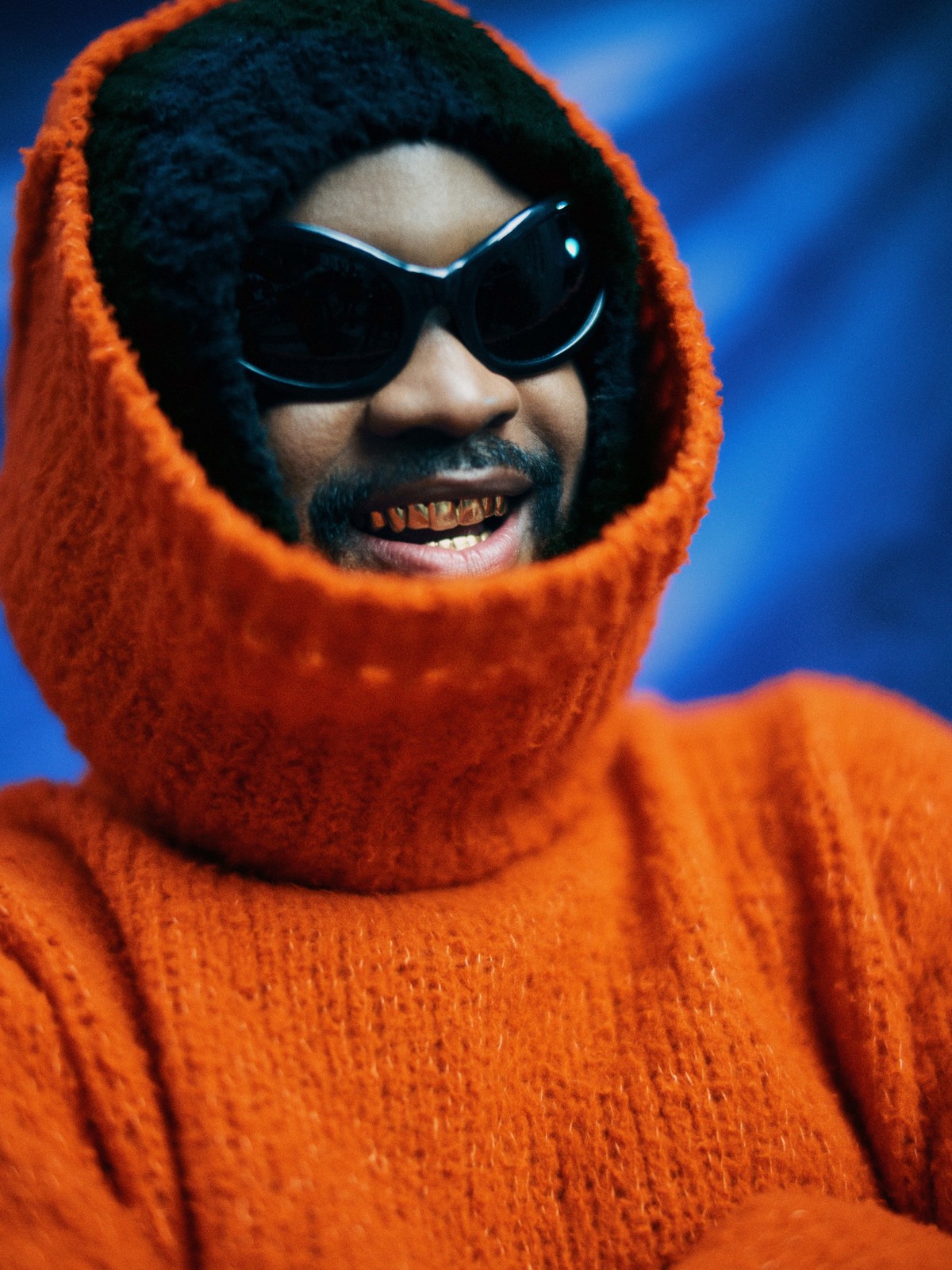 Close-up studio shot of an adult Black man with gold teeth wearing a bright orange hooded sweater and sunglasses against a blue background.