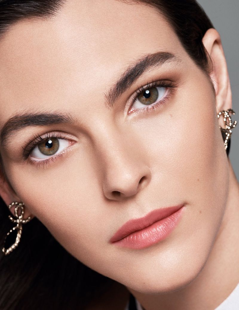 Close-up portrait of a woman with fair skin, light eyes, and soft makeup. She wears gold earrings and faces the camera directly.