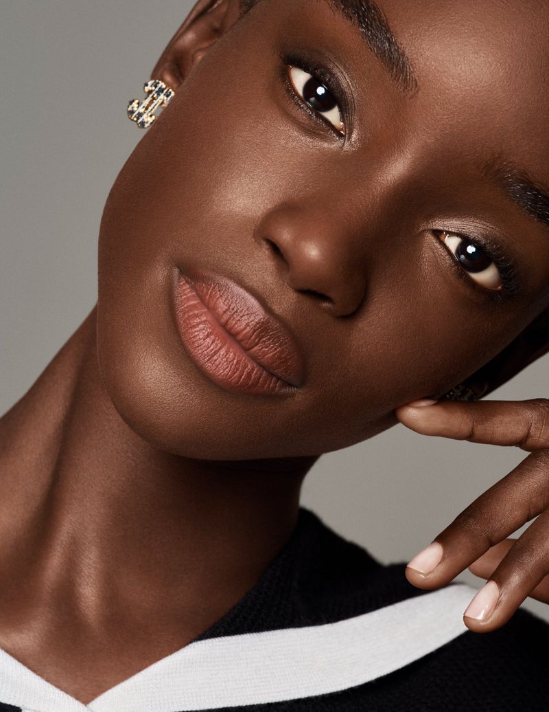 Close-up portrait of a dark-skinned Black woman with dark eyes and a neutral expression, wearing earrings and a black and white top in studio.
