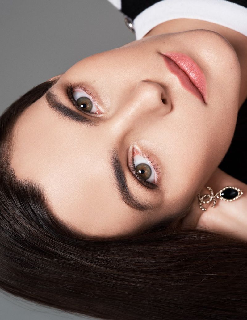 Close-up portrait of a woman with long brown hair, fair skin, and brown eyes looking at the camera against a grey studio backdrop.