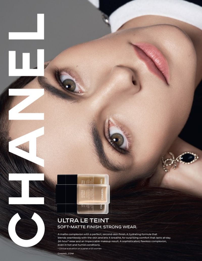 Close-up of an adult female model wearing makeup and jewelry, with CHANEL Ultra Le Teint foundation bottles in the foreground, studio lighting.