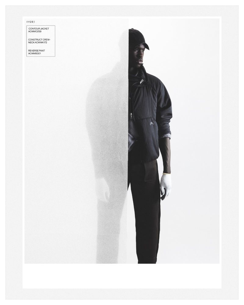Male fashion model in a dark jacket, black pants, and a cap, showcased in a studio setting with bright lighting. 