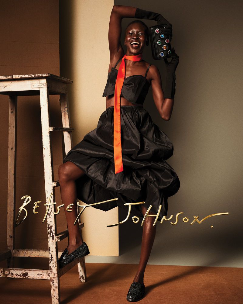 A Black fashion model wearing a dark two-piece outfit, gloves, and loafers is posing with a patterned purse and bright orange tie in a studio.