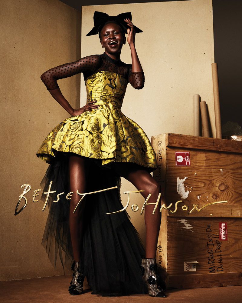 A Black woman in a yellow floral gown with a large black bow poses in front of wooden crates in a fashion editorial shot.