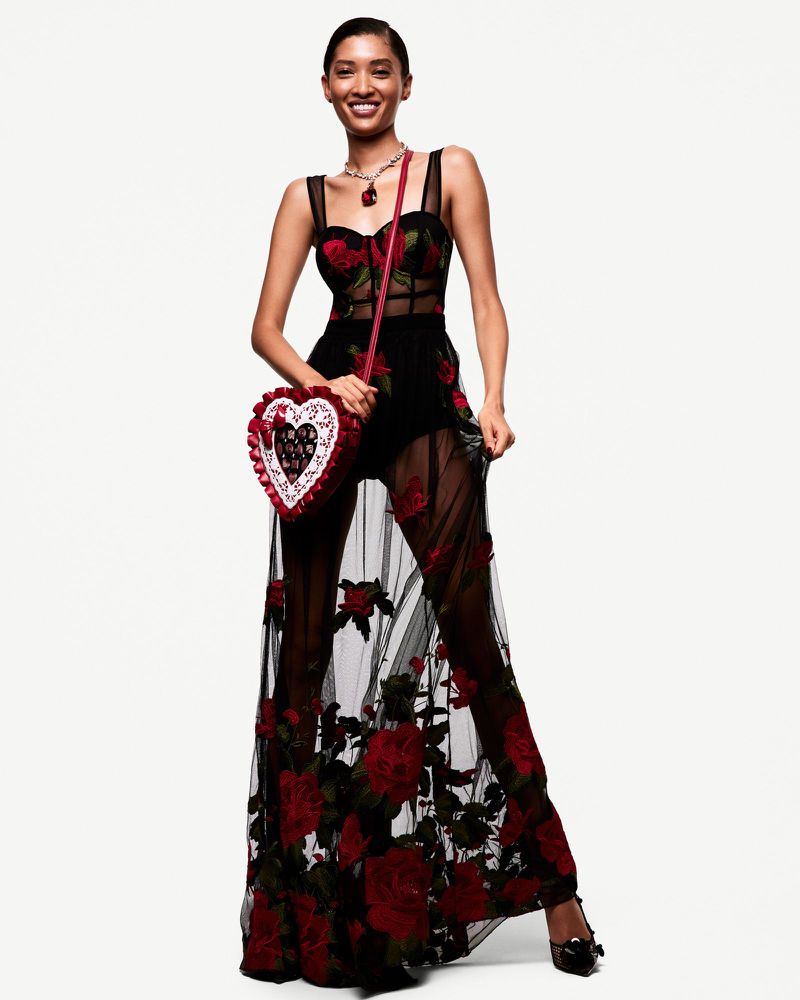 A striking fashion portrait of a smiling Black woman in a rose-embroidered gown, heart-shaped purse, and necklace against a clean white backdrop.