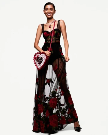 A striking fashion portrait of a smiling Black woman in a rose-embroidered gown, heart-shaped purse, and necklace against a clean white backdrop.