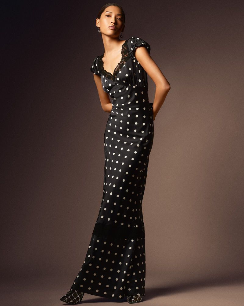 Model wearing a black polka-dot maxi dress with lace trim standing against a seamless dark studio background
