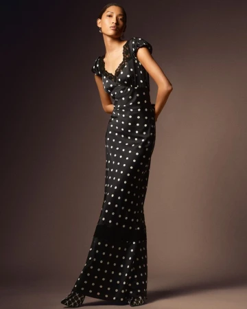 Model wearing a black polka-dot maxi dress with lace trim standing against a seamless dark studio background