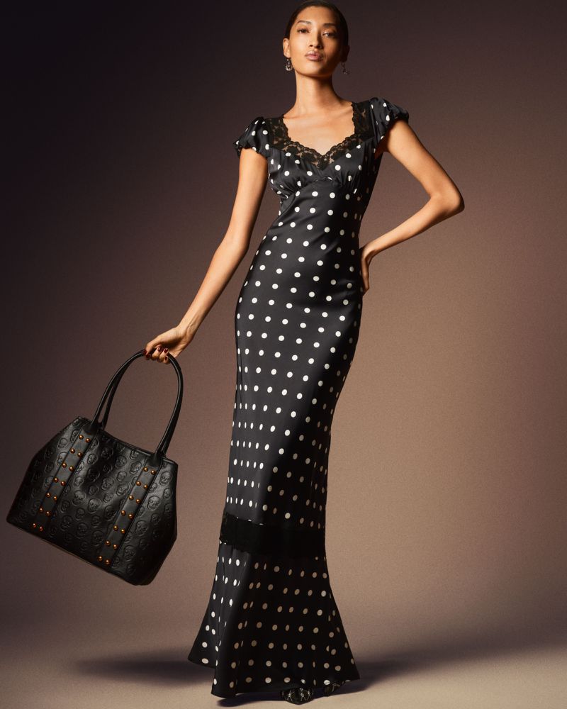 Elegant adult woman in a black-and-white polka dot dress holding a black leather handbag, posing in a photo studio with neutral backdrop.