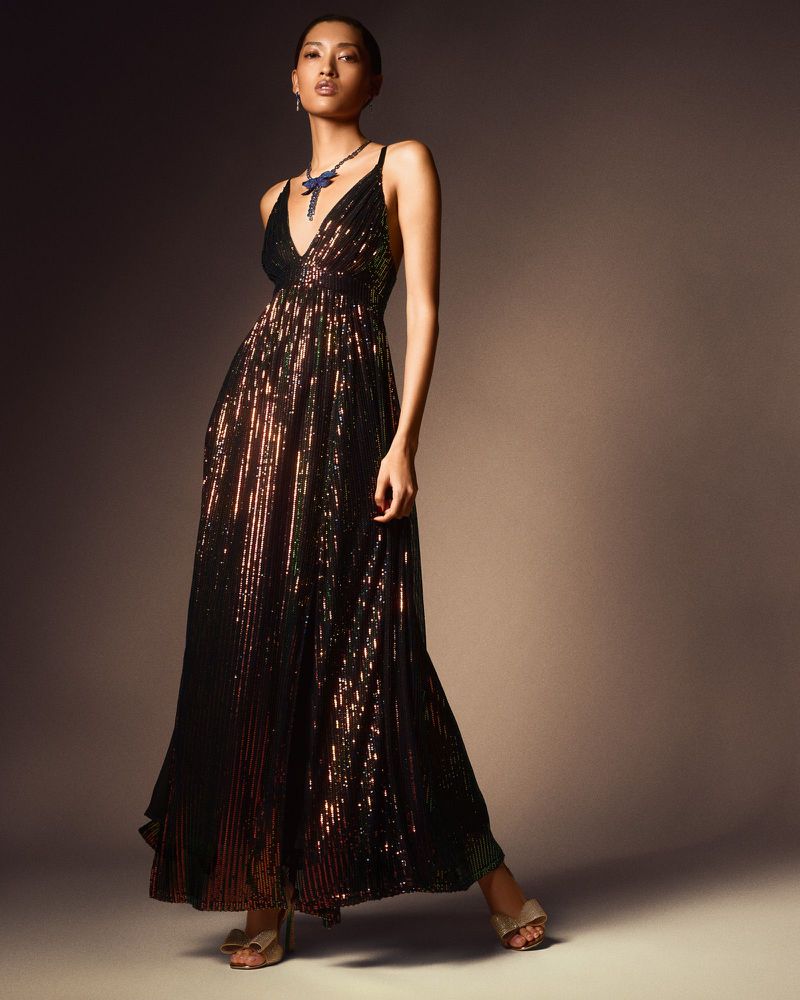 Elegant woman in a shimmering gown with a V-neckline and delicate necklace, posing confidently against a neutral backdrop in a fashion shot.