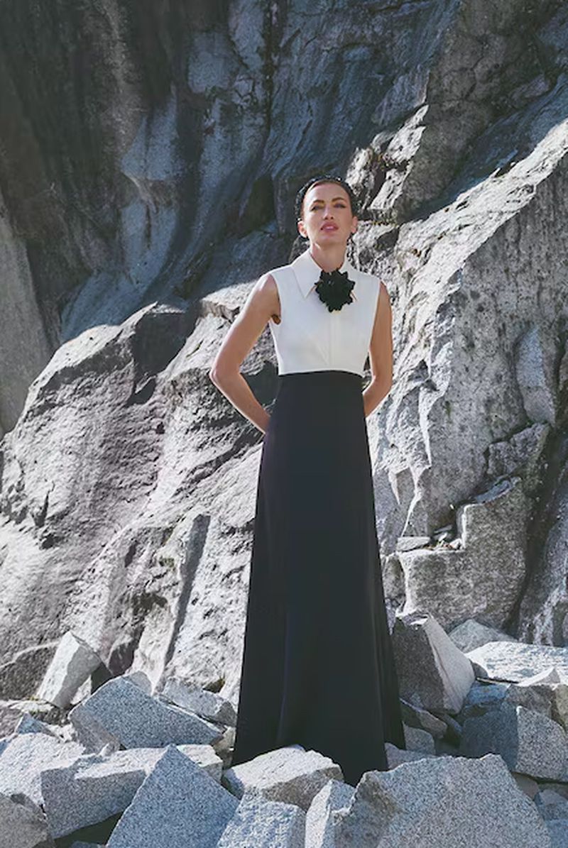 A fashion photo of a woman in a black and white dress posing outdoors on a sunny day with a large rock formation in the background.