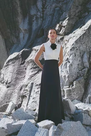 A fashion photo of a woman in a black and white dress posing outdoors on a sunny day with a large rock formation in the background.