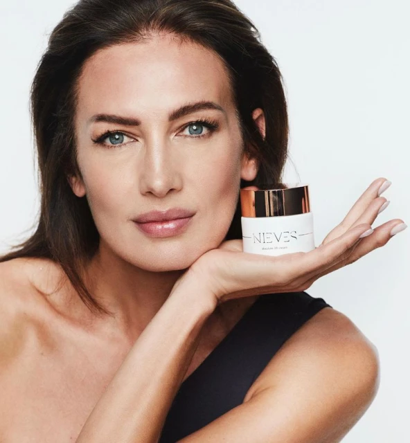 Adult female model poses with a white cosmetic jar with gold accents in a close-up studio shot against a bright white background.