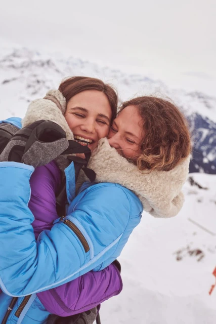Two young adult women embrace warmly in a snowy mountain environment, smiling and bundled in winter clothing and gear, showing joy and affection.