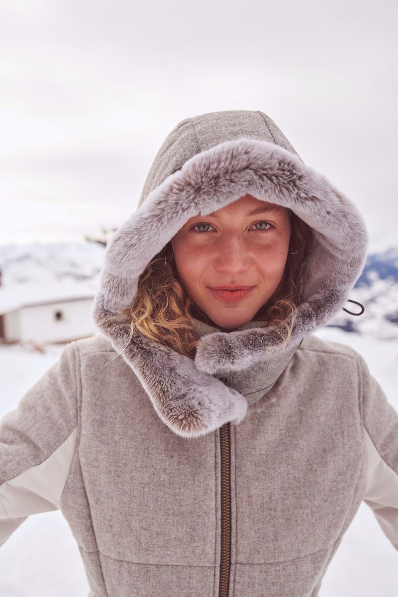 Smiling woman in a winter jacket with fur-lined hood stands in a snowy landscape. Close-up portrait in natural light with a neutral expression.