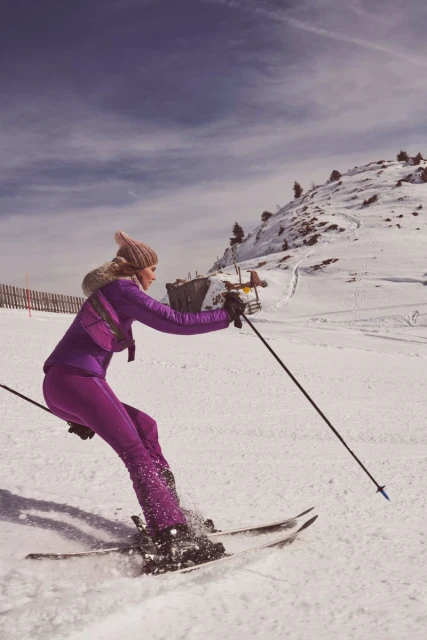 A woman in a purple ski suit skis down a snow-covered mountain, with the sky visible behind her. She holds ski poles as she moves.