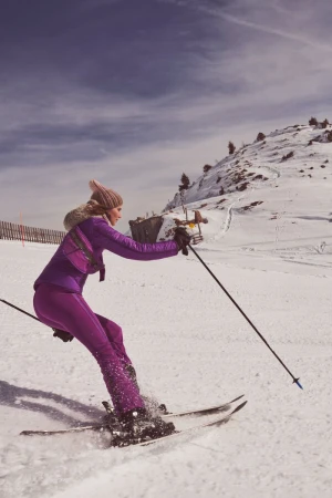 A woman in a purple ski suit skis down a snow-covered mountain, with the sky visible behind her. She holds ski poles as she moves.
