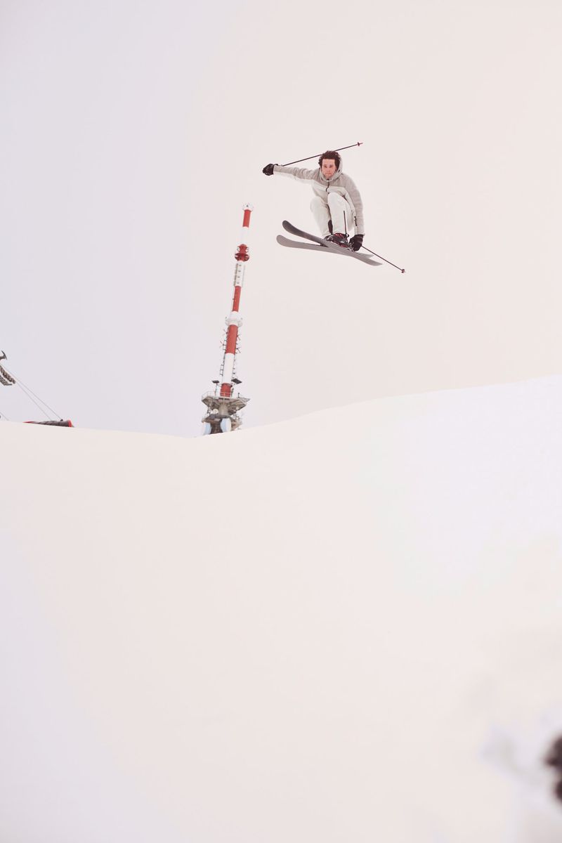 Male skier performs mid-air jump near a red and white pole on snowy terrain. Action shot captures a winter sports moment in bright daylight.