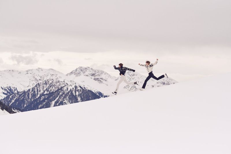 Two adults leap across a snow-covered mountain landscape on a cloudy day, showcasing winter fun, athleticism, and scenic mountain views.