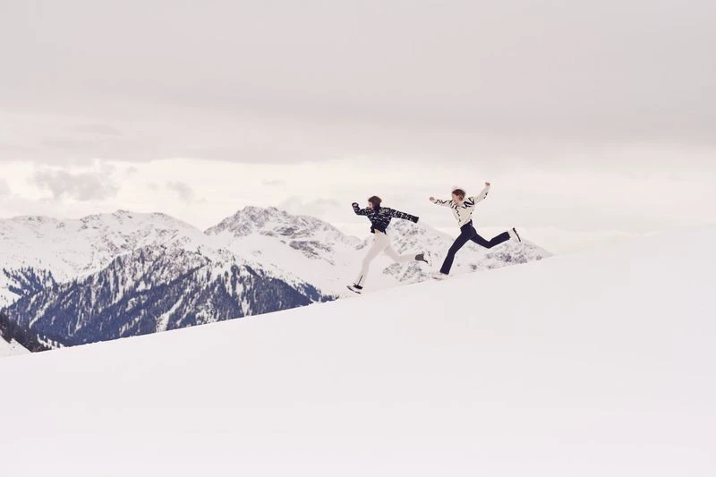 Two adults leap across a snow-covered mountain landscape on a cloudy day, showcasing winter fun, athleticism, and scenic mountain views.