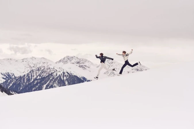 Two adults leap across a snow-covered mountain landscape on a cloudy day, showcasing winter fun, athleticism, and scenic mountain views.